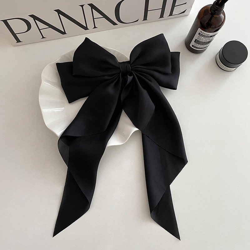 Barrete noeud papillon noir en satin pour les cheveux posée sur une assiette blanche entourée de deux produits de beauté et d'un sac "panache"